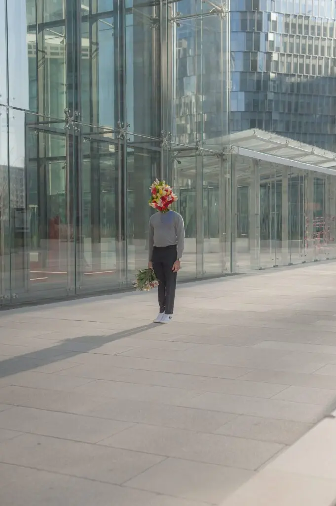 Conceptual photograph of a person in a gray sweater standing outside a modern glass building with a bouquet of flowers covering their head, holding another bouquet in hand.