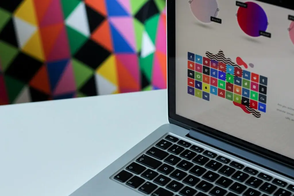 Close-up of a laptop screen displaying colorful design icons and palettes, with a vibrant geometric wall pattern in the background.