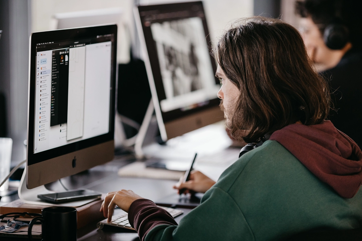 Person with long hair wearing a green hoodie works at a desktop computer in a shared office, using a drawing tablet and pen