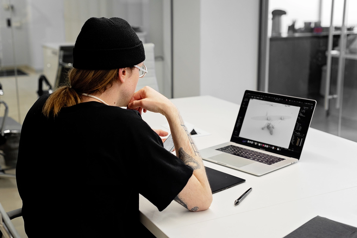 Person with a black beanie and glasses sits at a white desk, looking at a laptop displaying a grayscale 3D drone model.