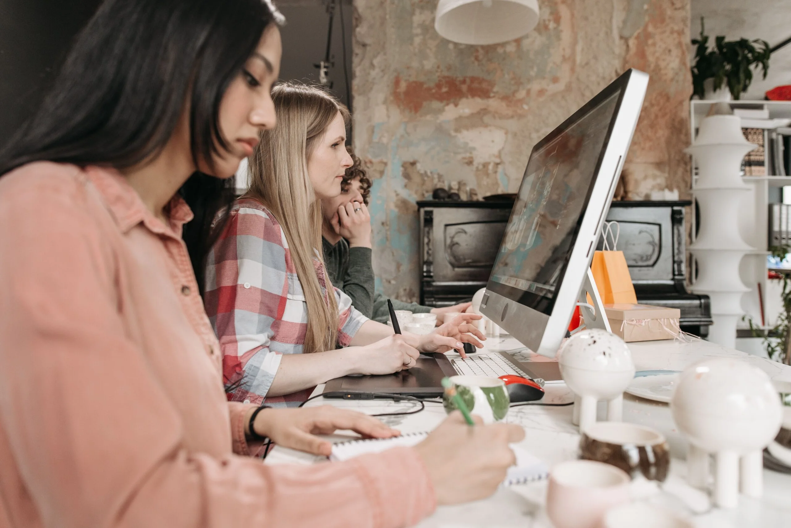 Group of people working at a bright designer studio, focusing on a large computer screen and sketching at the desk.
